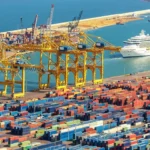 A bustling container port with colorful shipping containers stacked neatly under yellow gantry cranes. A large cruise ship sails nearby, representing global connectivity. This scene reflects the export strength of Yiwu’s small commodity market and the role of platforms like yiwuagents.com in facilitating international sourcing and cross-border trade.