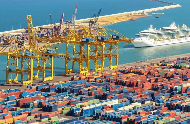 A bustling container port with colorful shipping containers stacked neatly under yellow gantry cranes. A large cruise ship sails nearby, representing global connectivity. This scene reflects the export strength of Yiwu’s small commodity market and the role of platforms like yiwuagents.com in facilitating international sourcing and cross-border trade.